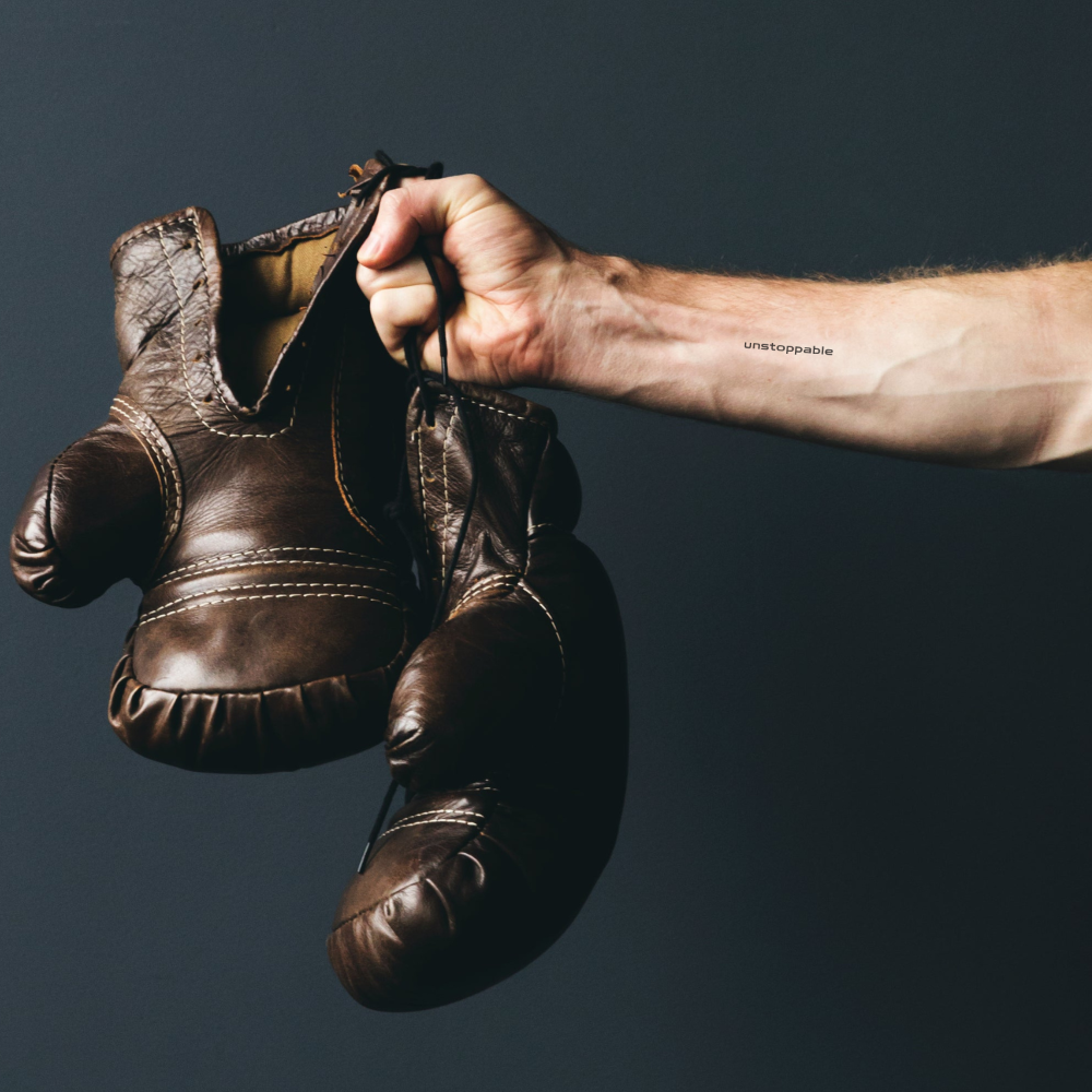 'unstoppable' empowering temporary tattoo on inner forearm – minimalist black ink fitness quote by Motivink with vintage boxing gloves