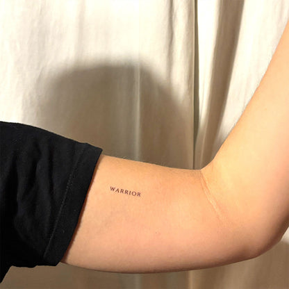 Black ink ‘WARRIOR’ motivational temporary tattoo on inner arm – minimalist quote design for strength, self-belief, and empowerment by MotivInk
